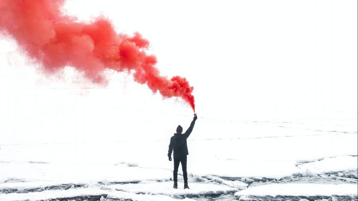 A person standing in a middle of a small hilly field with red smoke coming off their hand in the middle of their air.