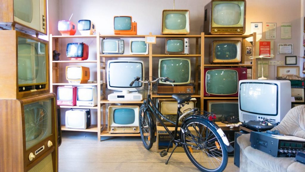 A room with several TVs from the 70s to 90s on several shelves. A bike and couch are also in this storage room.
