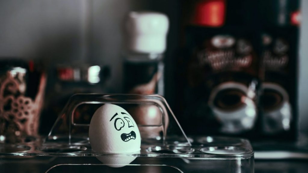 An egg resting on a clear glass holder with a inked facial expression of frustration. The background is a kitchen counter that has been blurred.