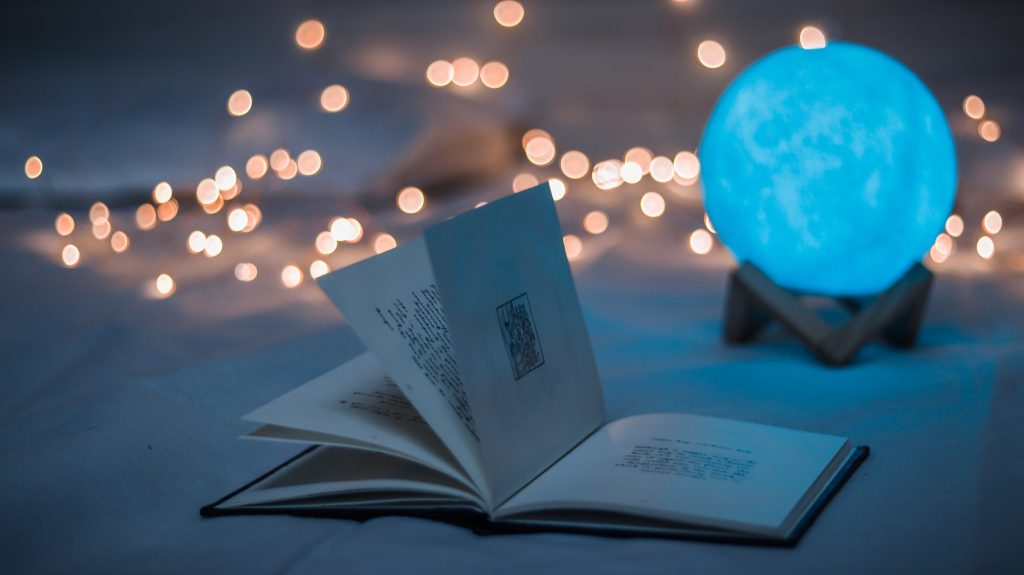 A book magically opening without any aid sitting in front a light blue glowing ball. Sparkling of lights lean on the background.